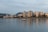 High-rise buildings along a waterfront with calm waters reflecting the structures. A small boat is in the foreground, and mountains are visible in the distance under an overcast sky.