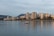 High-rise buildings along a waterfront with calm waters reflecting the structures. A small boat is in the foreground, and mountains are visible in the distance under an overcast sky.