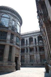 A historic and grand architectural courtyard is flanked by tall stone buildings, featuring multiple floors and large, intricate windows. The design demonstrates classical architectural elements such as columns and arched windows.