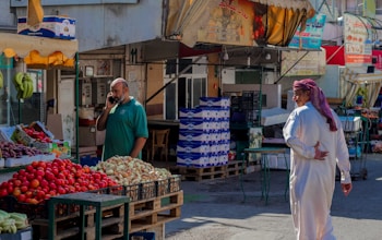 A market scene with a man talking on a phone near a fruit stand displaying apples, bananas, and onions. Another man in traditional clothing walks by. There are signs in Arabic above multiple stalls, and the area is shaded by awnings.