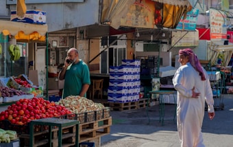 A market scene with a man talking on a phone near a fruit stand displaying apples, bananas, and onions. Another man in traditional clothing walks by. There are signs in Arabic above multiple stalls, and the area is shaded by awnings.
