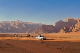 A desert landscape with a rugged mountain range in the background and a sandy terrain in the foreground. A group of people riding camels is seen traveling alongside a white vehicle parked on the sand.