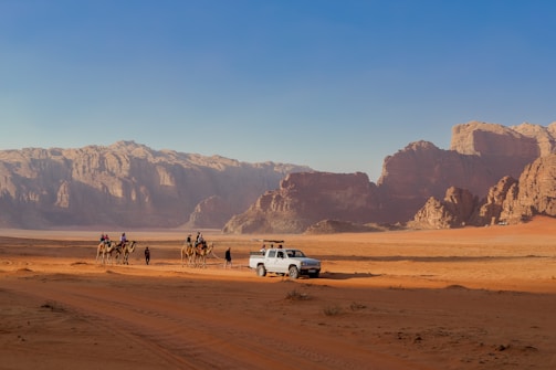 A desert landscape with a rugged mountain range in the background and a sandy terrain in the foreground. A group of people riding camels is seen traveling alongside a white vehicle parked on the sand.