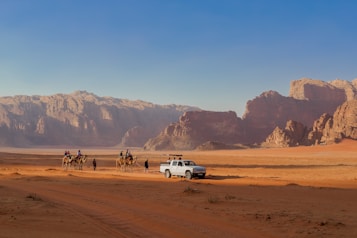 A desert landscape with a rugged mountain range in the background and a sandy terrain in the foreground. A group of people riding camels is seen traveling alongside a white vehicle parked on the sand.