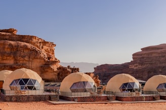 Geodesic dome structures made from beige material are set against a backdrop of striking sandstone cliffs and a clear blue sky. The domes have triangular window panels, suggesting they might be modern tents or lodging accommodations. The scene has a desert-like setting with sand covering the ground.