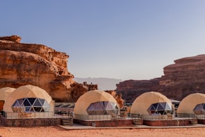 Geodesic dome structures made from beige material are set against a backdrop of striking sandstone cliffs and a clear blue sky. The domes have triangular window panels, suggesting they might be modern tents or lodging accommodations. The scene has a desert-like setting with sand covering the ground.
