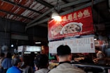 An indoor market scene features a group of people gathered around a food stall. The stall is adorned with brightly colored signage in Chinese, displaying images of dishes. The setting appears busy, with patrons waiting in line and a vendor serving food.