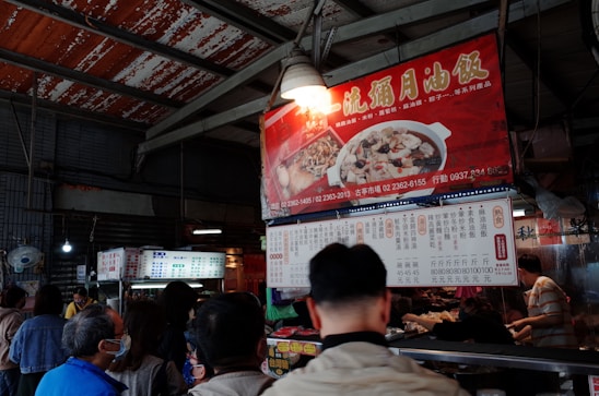 An indoor market scene features a group of people gathered around a food stall. The stall is adorned with brightly colored signage in Chinese, displaying images of dishes. The setting appears busy, with patrons waiting in line and a vendor serving food.