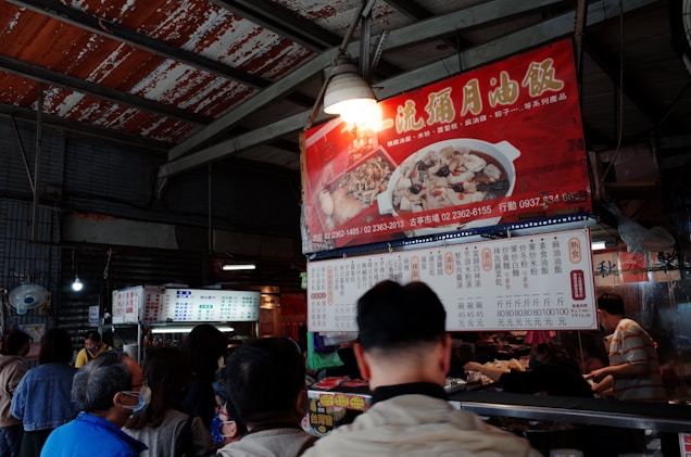 An indoor market scene features a group of people gathered around a food stall. The stall is adorned with brightly colored signage in Chinese, displaying images of dishes. The setting appears busy, with patrons waiting in line and a vendor serving food.