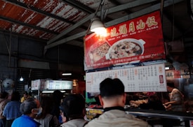 An indoor market scene features a group of people gathered around a food stall. The stall is adorned with brightly colored signage in Chinese, displaying images of dishes. The setting appears busy, with patrons waiting in line and a vendor serving food.