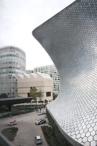 A modern architectural scene featuring a large, curved structure with a hexagonal patterned facade, an adjacent multi-story circular building, and a square contemporary building in the background. The area has a courtyard with several cars parked, people walking, and landscaped greenery.