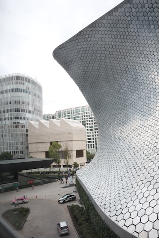 A modern architectural scene featuring a large, curved structure with a hexagonal patterned facade, an adjacent multi-story circular building, and a square contemporary building in the background. The area has a courtyard with several cars parked, people walking, and landscaped greenery.