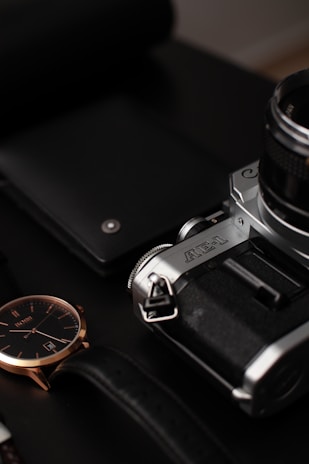 A vintage camera with a black body and silver accents is prominently displayed. Next to the camera lies a sleek black closed notebook. A stylish wristwatch with a black leather strap and a dark face sits nearby, exuding elegance with its gold or rose gold case.