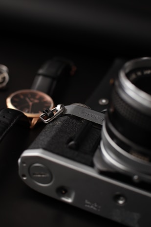 A close-up of a vintage camera alongside a wristwatch with a black leather strap and a gold casing. The image features a primarily dark background with a sense of elegance and sophistication.