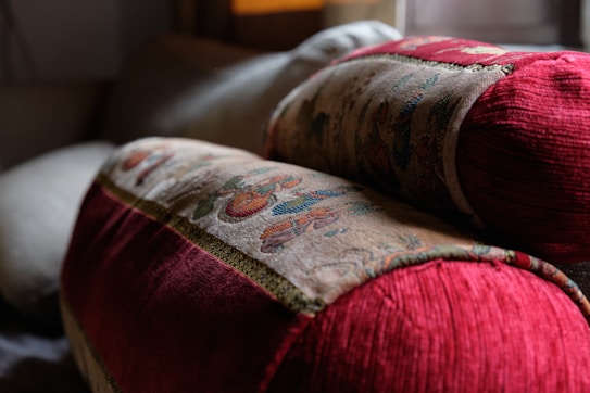 Two intricately patterned cushions with rich red fabric and detailed embroidery rest on a soft surface, creating a cozy and inviting atmosphere in what appears to be a living space with soft lighting.