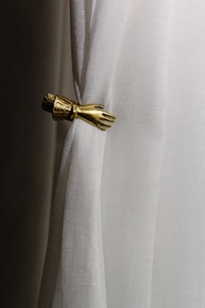 A white curtain is gathered by an ornate metallic hand-shaped tieback. The fabric has a slightly textured appearance, while the tieback is detailed with an elegant design resembling a hand and cuff.