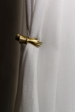 A white curtain is gathered by an ornate metallic hand-shaped tieback. The fabric has a slightly textured appearance, while the tieback is detailed with an elegant design resembling a hand and cuff.