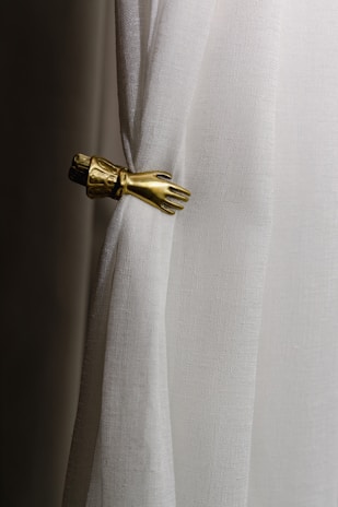 A white curtain is gathered by an ornate metallic hand-shaped tieback. The fabric has a slightly textured appearance, while the tieback is detailed with an elegant design resembling a hand and cuff.