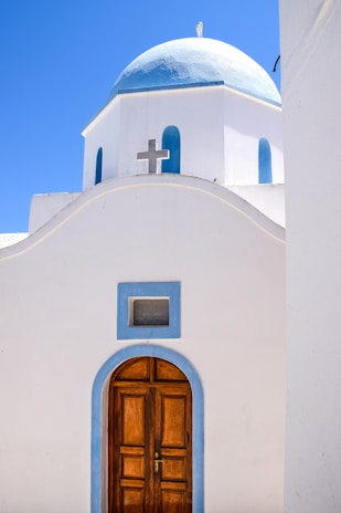 A white church building with a blue dome is set against a bright blue sky. The structure features a wooden arched door accented with a light blue frame, and a prominent cross is visible above an architectural curve.