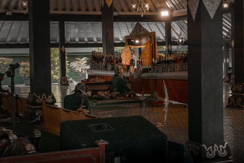 A traditional puppet show is taking place in a dimly lit structure with a high ceiling. Several people are sitting and watching the performance, which features intricately designed puppets and vibrant costumes. The setting includes traditional decorations and soft ambient lighting.