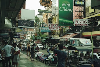 A busy urban street filled with various shop signs, people walking, and vehicles, including motorbikes and tuk-tuks. The atmosphere is lively, with colorful advertisements and buildings crowding the scene. Many individuals are visible, some standing while others are walking about.