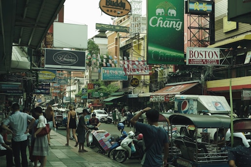 A busy urban street filled with various shop signs, people walking, and vehicles, including motorbikes and tuk-tuks. The atmosphere is lively, with colorful advertisements and buildings crowding the scene. Many individuals are visible, some standing while others are walking about.