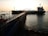 A large tanker ship is docked at a pier, which extends into the water. The structure is equipped with pipes and industrial equipment. The sun is setting in the background, casting a warm glow over the scene. The water reflects the light, creating a serene and industrial atmosphere.