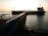 A large tanker ship is docked at a pier, which extends into the water. The structure is equipped with pipes and industrial equipment. The sun is setting in the background, casting a warm glow over the scene. The water reflects the light, creating a serene and industrial atmosphere.