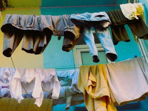 A collection of various clothing items such as pants, shirts, and towels hanging from a clothesline against a colorful wall. The scene is outdoors with a window visible in the background.