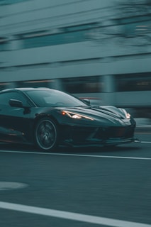 A sleek, black sports car is captured in motion against an urban backdrop. The vehicle's glossy finish reflects the muted city lights as it speeds down a wide road. The background appears slightly blurred, emphasizing the car's speed and dynamic presence.