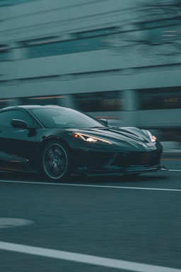 A sleek, black sports car is captured in motion against an urban backdrop. The vehicle's glossy finish reflects the muted city lights as it speeds down a wide road. The background appears slightly blurred, emphasizing the car's speed and dynamic presence.