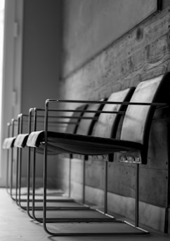 A row of modern, metal-framed chairs with dark seats and backs, lined up against a textured wooden wall. The chairs have a sleek, minimalist design and the light coming through the window casts soft shadows on the floor, enhancing the monochrome style of the scene.