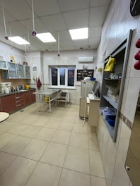 A spacious, well-lit kitchen with tiled floors and walls. There are wooden cabinets with glass doors on one side, and a countertop with various cooking appliances like a mixer. A table with two chairs is placed near the window. On the opposite side, there's a fridge with various items placed on top. The ceiling is lined with fluorescent lights.