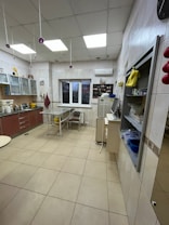 A spacious, well-lit kitchen with tiled floors and walls. There are wooden cabinets with glass doors on one side, and a countertop with various cooking appliances like a mixer. A table with two chairs is placed near the window. On the opposite side, there's a fridge with various items placed on top. The ceiling is lined with fluorescent lights.