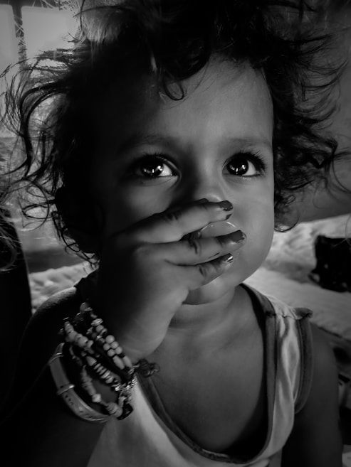 A young child with curly hair and expressive eyes covers their mouth with their hand. The child is wearing several beaded bracelets on the wrist and has a thoughtful, slightly curious expression. The image is in black and white, creating a dramatic and intimate feel.