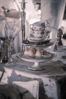 An elegantly set table features a tower of stacked teacups and saucers with silver detailing, alongside a serving of brown eggs on a pedestal dish. A tall silver candlestick and a silver champagne bucket with glasses add to the vintage ambiance.