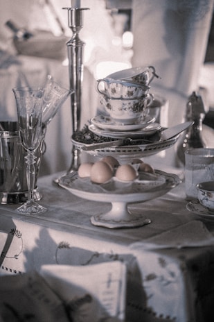 An elegantly set table features a tower of stacked teacups and saucers with silver detailing, alongside a serving of brown eggs on a pedestal dish. A tall silver candlestick and a silver champagne bucket with glasses add to the vintage ambiance.
