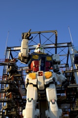 A giant humanoid robot figure stands against a clear blue sky, surrounded by a metal scaffolding structure. The robot has a vibrant color scheme with white, red, blue, and yellow sections and is positioned upright with one arm raised.