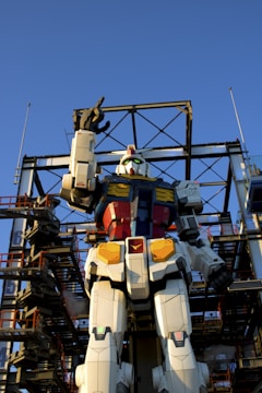 A giant humanoid robot figure stands against a clear blue sky, surrounded by a metal scaffolding structure. The robot has a vibrant color scheme with white, red, blue, and yellow sections and is positioned upright with one arm raised.