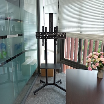 A modern office space with a glass wall and window blinds. There is a TV stand with wheels and a mounted bracket, placed in front of a window. A bouquet of pink and white flowers is arranged in a vase on a wooden desk.