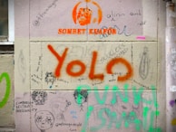 Graffiti covering a wall with various drawings, writings, and colorful text. Prominent words include 'YOLO' in large orange and 'SONBET KUAFOR' in smaller orange print. Other scribbles and drawings include faces and phrases like 'Comparison Destroys Personality' and '@myteasgxnecold - Lara.' The wall is divided into sections, each featuring different artistic expressions and comments.