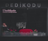 A man sits inside a dimly lit establishment with the word 'Dedikodu' written in large letters on the exterior. He is partially obscured by reflections on the window, appears to be deep in thought with his hand on his chin. Bright red seats and a few potted plants with pink flowers add contrast to the otherwise dark surroundings.