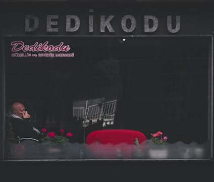 A man sits inside a dimly lit establishment with the word 'Dedikodu' written in large letters on the exterior. He is partially obscured by reflections on the window, appears to be deep in thought with his hand on his chin. Bright red seats and a few potted plants with pink flowers add contrast to the otherwise dark surroundings.
