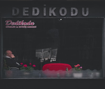 A man sits inside a dimly lit establishment with the word 'Dedikodu' written in large letters on the exterior. He is partially obscured by reflections on the window, appears to be deep in thought with his hand on his chin. Bright red seats and a few potted plants with pink flowers add contrast to the otherwise dark surroundings.