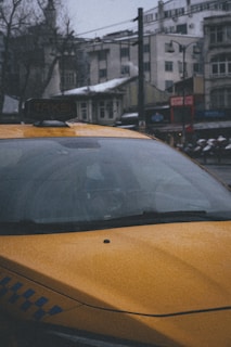 A yellow taxi is parked in an urban setting with snow visible on the rooftops and cars. The scene is slightly overcast and the buildings in the background appear to be older, with a combination of residential and commercial structures.
