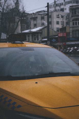 A yellow taxi is parked in an urban setting with snow visible on the rooftops and cars. The scene is slightly overcast and the buildings in the background appear to be older, with a combination of residential and commercial structures.