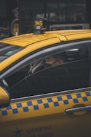 A person is seated inside a yellow taxi, with the focus on the vehicle's distinctive blue checkered pattern on the side and the illuminated 'TAKSI' sign on the roof. The person is wearing a face mask, looking out of the partially open window in an urban environment.