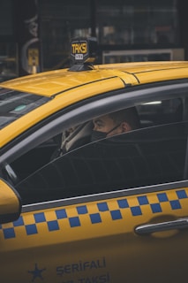 A person is seated inside a yellow taxi, with the focus on the vehicle's distinctive blue checkered pattern on the side and the illuminated 'TAKSI' sign on the roof. The person is wearing a face mask, looking out of the partially open window in an urban environment.