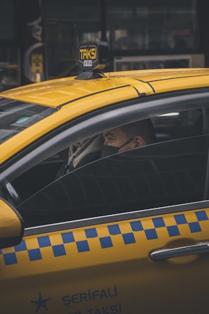 A person is seated inside a yellow taxi, with the focus on the vehicle's distinctive blue checkered pattern on the side and the illuminated 'TAKSI' sign on the roof. The person is wearing a face mask, looking out of the partially open window in an urban environment.