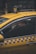 A person is seated inside a yellow taxi, with the focus on the vehicle's distinctive blue checkered pattern on the side and the illuminated 'TAKSI' sign on the roof. The person is wearing a face mask, looking out of the partially open window in an urban environment.